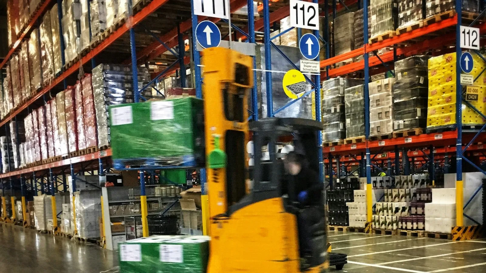 A reach truck carrying a pallet down a warehouse aisle between tall racks (stock photo)